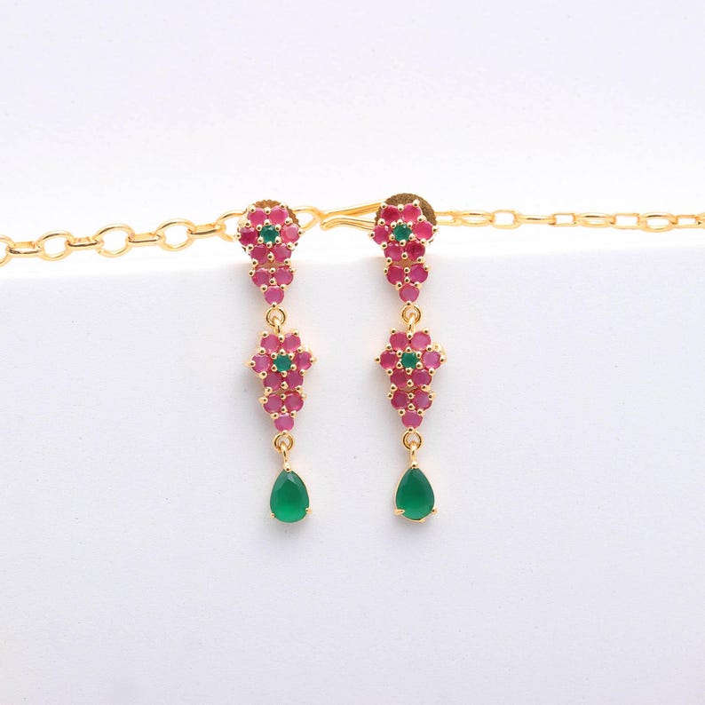 May include: A pair of gold-toned dangle earrings. Each earring features a cluster of small, pink gemstones with a central green gemstone. A teardrop-shaped green gemstone hangs from each earring.