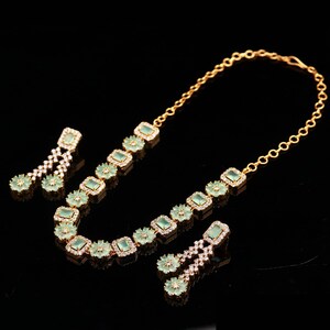 May include: A gold-toned jewelry set featuring a necklace and two pairs of earrings. The necklace and earrings are adorned with square, light green gemstones and small, clear crystals. The set is displayed against a black background.