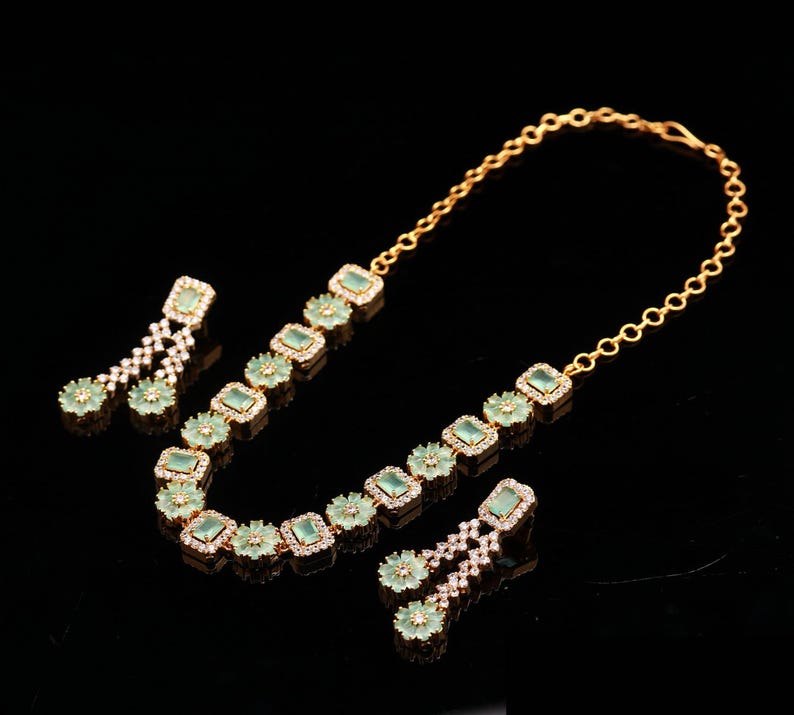 May include: A gold-toned jewelry set featuring a necklace and two pairs of earrings. The necklace and earrings are adorned with square, light green gemstones and small, clear crystals. The set is displayed against a black background.