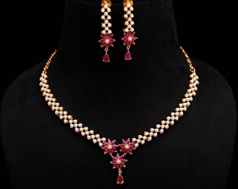 May include: A gold-toned necklace and earring set. The necklace features a double strand of small white beads with ruby-colored floral accents and a teardrop pendant. The matching earrings have a similar design, with ruby-colored floral accents and teardrop pendants.