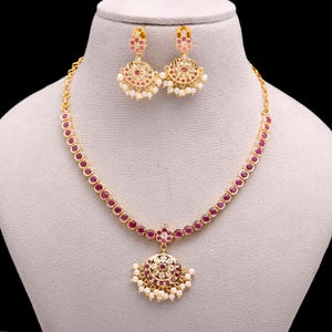 Op de afbeelding: Een gouden ketting en oorbellen set op een mannequin. De ketting heeft rode edelstenen en een hanger met een cirkelvormig ontwerp. De oorbellen hebben hetzelfde ontwerp, met rode edelstenen en witte parels. Voor speciale gelegenheden.
