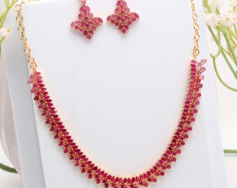 May include: A gold-toned necklace and earring set. The necklace and earrings are adorned with numerous small, oval-shaped, ruby-colored stones. The earrings are a diamond-like shape. The necklace has a curved design, with the stones set closely together.