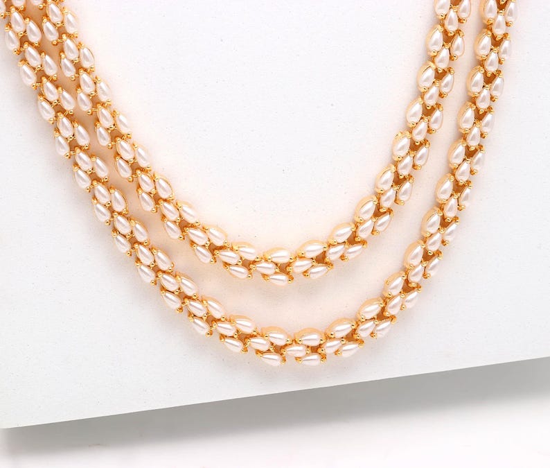 May include: Three-strand necklace featuring creamy white, teardrop-shaped beads. The beads are strung together with gold-colored metal links. The necklace is displayed on a white surface, showcasing its elegant design.