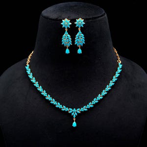 May include: A gold-toned jewelry set featuring a necklace and matching earrings. The necklace and earrings are adorned with turquoise-colored gemstones arranged in a leaf-like pattern. The earrings have a floral design at the top.