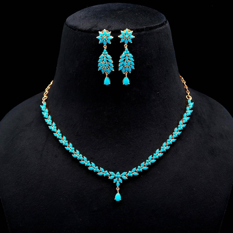 May include: A gold-toned jewelry set featuring a necklace and matching earrings. The necklace and earrings are adorned with turquoise-colored gemstones arranged in a leaf-like pattern. The earrings have a floral design at the top.