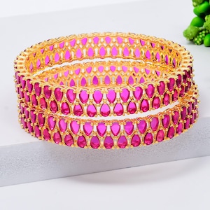 May include: Two gold bangles, each adorned with a repeating pattern of pink, teardrop-shaped stones. The bangles are stacked, highlighting the intricate design and vibrant colour of the jewellery. The background is a soft white.