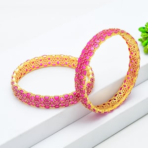 May include: Two gold bangles with a vibrant pink gemstone design. The gemstones are oval-shaped and closely set, creating a continuous pattern around the bangles. The gold setting has an openwork design.