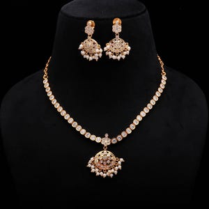 Puede incluir: Un conjunto de joyas dorado que incluye un collar y pendientes a juego. El collar presenta piedras redondas y transparentes y un colgante con un diseño floral y pequeñas perlas blancas. Los pendientes tienen un diseño similar.