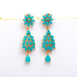 May include: A pair of gold-toned earrings featuring turquoise-colored stones. Each earring has a floral top, a cluster of stones, and a teardrop-shaped stone at the bottom. The earrings are set against a white background.
