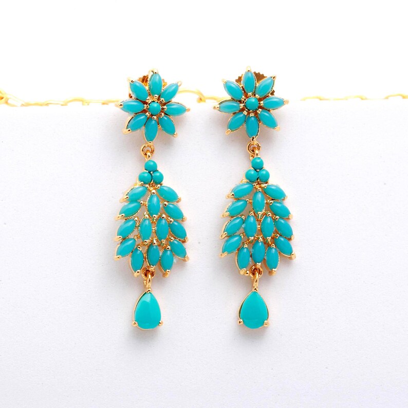 May include: A pair of gold-toned earrings featuring turquoise-colored stones. Each earring has a floral top, a cluster of stones, and a teardrop-shaped stone at the bottom. The earrings are set against a white background.