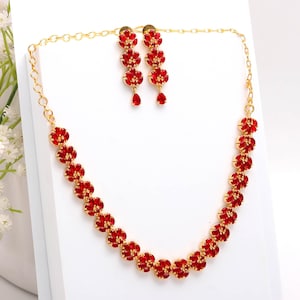 Puede incluir: Un conjunto de collar y pendientes de color dorado. El collar y los pendientes presentan un diseño floral con piedras preciosas rojas. Los pendientes son de estilo colgante, y el collar es de estilo gargantilla. Las joyas se exhiben sobre una superficie blanca.