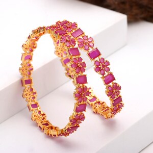 May include: Two gold-colored bangles with a floral design. Each bangle features alternating rectangular and flower-shaped magenta-colored gemstones. The bangles are displayed on a white surface.