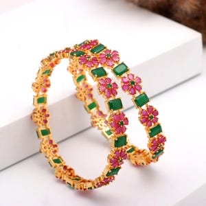 Peut inclure: Deux bracelets dorés ornés de motifs alternés de fleurs roses et de pierres vertes rectangulaires. Les bracelets sont présentés sur une surface blanche, mettant en valeur leurs motifs floraux et de pierres précieuses. La bijouterie est dorée.