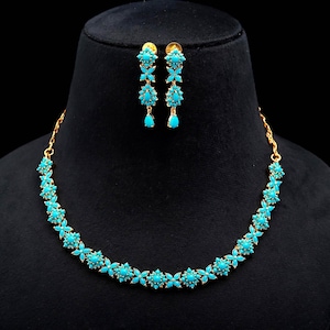 Può includere: Un set di collana e orecchini dorati con motivi floreali turchesi. La collana e gli orecchini sono esposti su una testa di manichino nera. Gli orecchini sono lunghi e pendono, mentre la collana è intorno al collo.