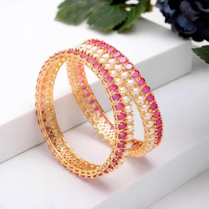 May include: Two gold bangles with alternating rows of pink and white gemstones. The bangles have a detailed, openwork design and are displayed on a white surface. The gemstones are oval-shaped.