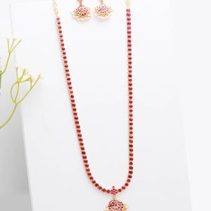 Pode incluir: Um conjunto de joias douradas com um colar longo e brincos a condizer. O colar e os brincos são adornados com pequenas pedras preciosas vermelhas redondas. O colar tem um pendente com um aglomerado de pedras vermelhas e pérolas brancas.