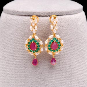 May include: A pair of gold-toned earrings featuring a central teardrop-shaped ruby surrounded by green and white gemstones. The earrings have a dangling ruby teardrop and a cluster of white pearls at the top.