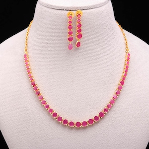May include: A gold-toned necklace and earring set. The necklace and earrings feature a series of round, pink gemstones. The earrings are dangling, with a teardrop-shaped gemstone at the bottom. The jewelry is displayed on a white mannequin.