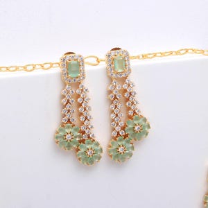May include: A gold-toned jewelry set featuring a necklace and earrings. The earrings have a square, light green stone at the top, followed by a cascade of clear stones and a flower-shaped pendant. The necklace has a similar design, with a square green stone and flower pendant.