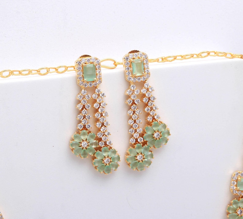 May include: A gold-toned jewelry set featuring a necklace and earrings. The earrings have a square, light green stone at the top, followed by a cascade of clear stones and a flower-shaped pendant. The necklace has a similar design, with a square green stone and flower pendant.