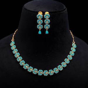 Könnte beinhalten: Ein goldfarbenes Halsketten- und Ohrring-Set. Die Halskette und die Ohrringe weisen ein Blumenmuster mit türkisfarbenen Steinen auf. Die Ohrringe sind hängend und die Halskette ist im Choker-Stil. Der Schmuck wird auf einer schwarzen Schaufensterpuppe präsentiert.