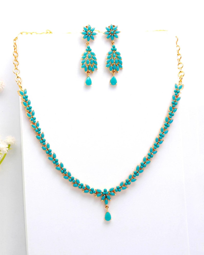 May include: A gold-toned jewelry set featuring a necklace and matching earrings. The necklace and earrings are adorned with turquoise-colored stones. The necklace has a floral design with a teardrop pendant. The earrings have a floral top and a teardrop bottom.