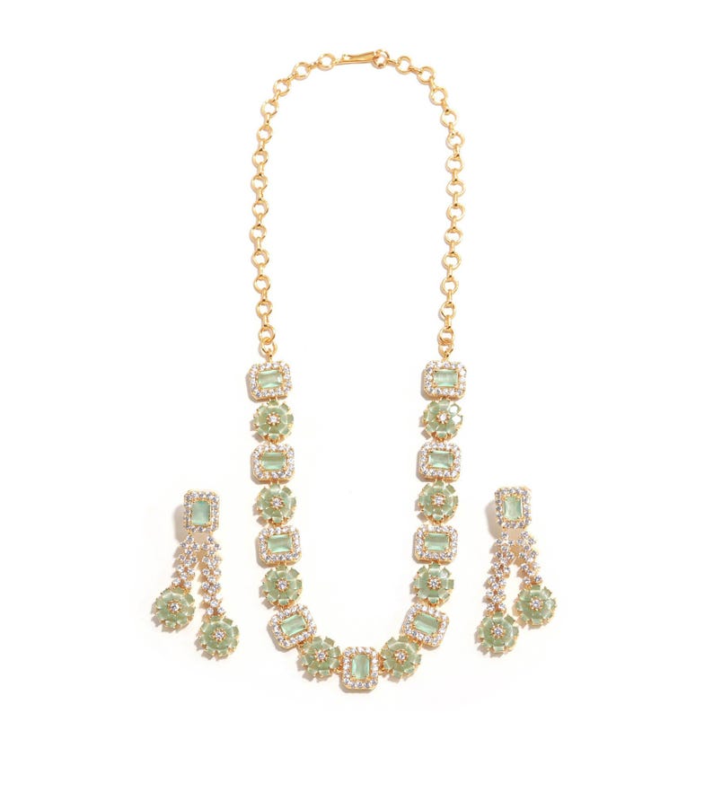 May include: A gold-toned necklace and matching earrings set. The necklace features rectangular light green stones surrounded by clear crystals. The earrings have a similar design, with dangling elements. The jewelry is set against a white background.