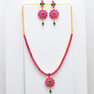May include: A gold-tone necklace and matching earring set featuring vibrant pink circular stones arranged in floral patterns, accented with small green stones and a single teardrop-shaped green stone pendant on the necklace and earrings.