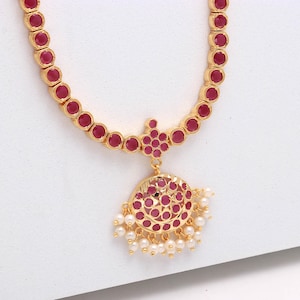 Könnte beinhalten: Goldfarbene Halskette mit einer Reihe runder, roter Edelsteine. Der Anhänger hat ein kreisförmiges Design mit weiteren roten Edelsteinen und kleinen weißen Perlen. Die Halskette wird auf einer weißen Oberfläche präsentiert.