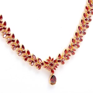 Puede incluir: Un collar dorado con un diseño floral, con numerosas piedras preciosas de color rojo intenso. El collar tiene un colgante en forma de lágrima en el centro. Las piedras están dispuestas en un delicado patrón en forma de hoja.