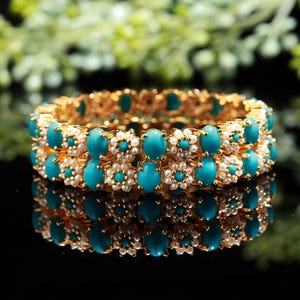 Pode incluir: Duas pulseiras douradas com pedras ovais turquesas e pequenos detalhes de p&eacute;rolas brancas. As pulseiras est&atilde;o empilhadas, refletindo em uma superf&iacute;cie preta. O fundo &eacute; folhagem verde desfocada.