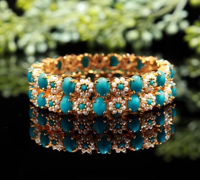 Pode incluir: Duas pulseiras douradas com pedras ovais turquesas e pequenos detalhes de p&eacute;rolas brancas. As pulseiras est&atilde;o empilhadas, refletindo em uma superf&iacute;cie preta. O fundo &eacute; folhagem verde desfocada.