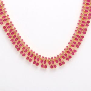 Peut inclure: Un collier doré orné d'une série de pierres précieuses ovales de couleur rubis. Les pierres sont disposées de près, créant un motif continu et scintillant. Le collier est présenté sur un fond blanc.
