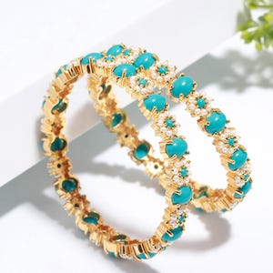 Pode incluir: Duas pulseiras douradas com pedras ovais turquesa e pequenos detalhes de p&eacute;rolas brancas. As pulseiras est&atilde;o sobre uma superf&iacute;cie branca. A joia &eacute; um acess&oacute;rio brilhante e chamativo.
