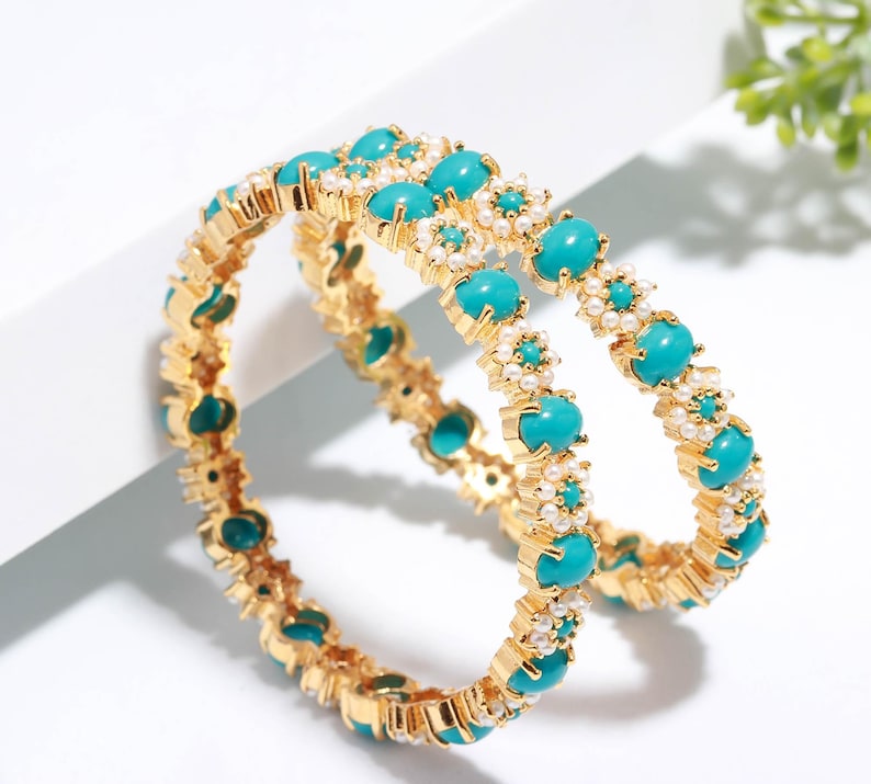 Pode incluir: Duas pulseiras douradas com pedras ovais turquesa e pequenos detalhes de p&eacute;rolas brancas. As pulseiras est&atilde;o sobre uma superf&iacute;cie branca. A joia &eacute; um acess&oacute;rio brilhante e chamativo.