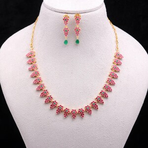 May include: A gold-toned necklace and earring set. The necklace and earrings feature a repeating pattern of pink and green gemstones. The earrings have a dangling green gemstone. The jewelry is displayed on a white mannequin.