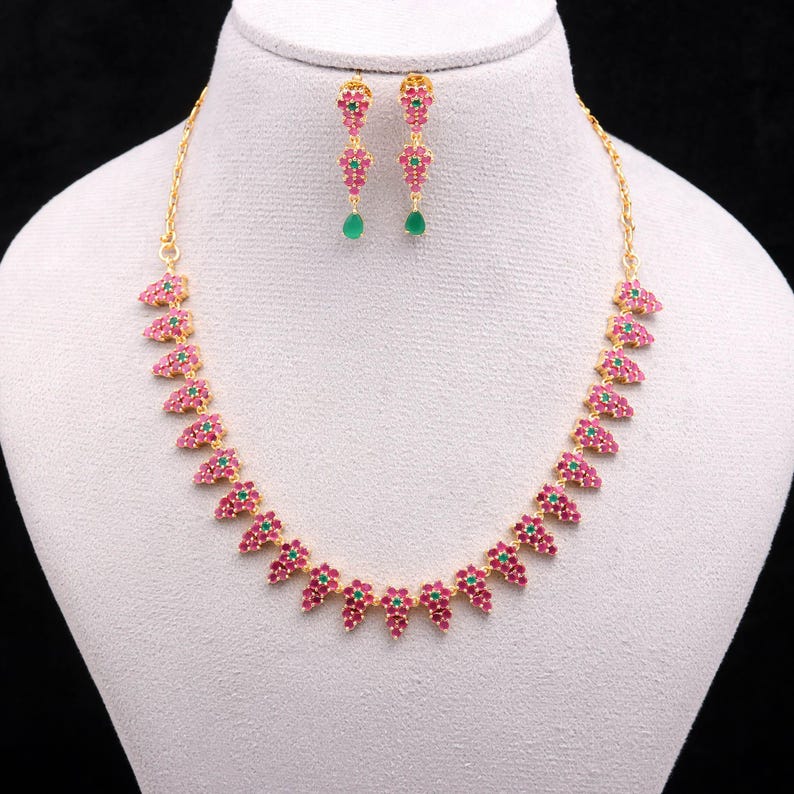 May include: A gold-toned necklace and earring set. The necklace and earrings feature a repeating pattern of pink and green gemstones. The earrings have a dangling green gemstone. The jewelry is displayed on a white mannequin.