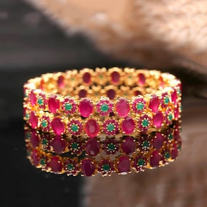 Könnte beinhalten: Goldfarbenes Armreif-Armband mit drei Reihen ovaler, rubinroter Edelsteine und kleinen, grünen Edelsteinen. Das Armband ist stark reflektierend, mit dunklem Hintergrund.