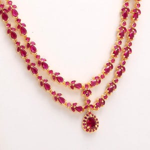 Peut inclure: Un collier en or à deux rangs orné de pierres précieuses de couleur rubis. Le collier comporte un pendentif en forme de goutte d'eau en bas et est posé sur un fond blanc. Les pierres précieuses sont d'un rouge profond.