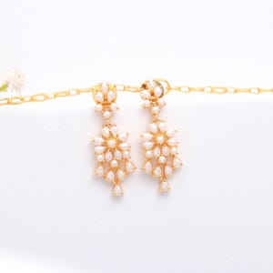 May include: A pair of gold-toned chandelier earrings with a floral design. Each earring features a cluster of small, white, teardrop-shaped pearls. The earrings are suspended from a gold chain, creating a delicate and elegant look.