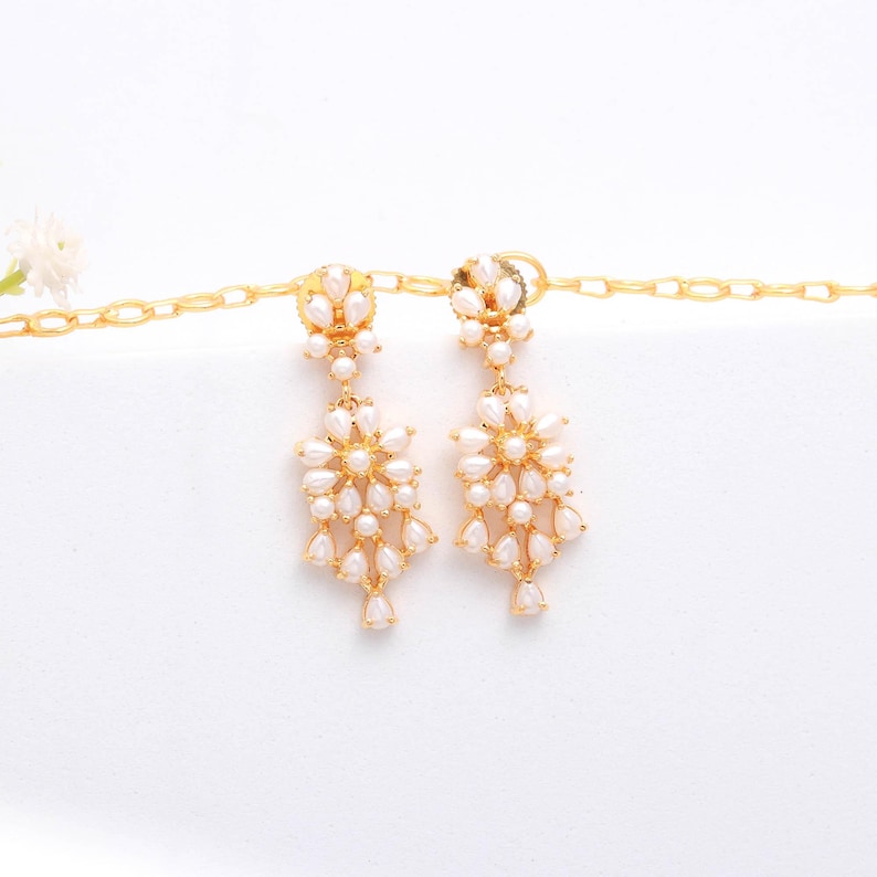 May include: A pair of gold-toned chandelier earrings with a floral design. Each earring features a cluster of small, white, teardrop-shaped pearls. The earrings are suspended from a gold chain, creating a delicate and elegant look.