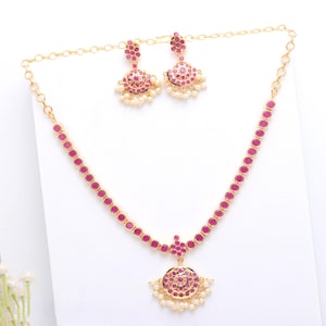 Puede incluir: Un conjunto de joyas de color dorado con un collar y pendientes a juego. El collar y los pendientes están adornados con piedras preciosas rosas redondas y pequeñas perlas blancas. El collar tiene un colgante con un diseño circular.