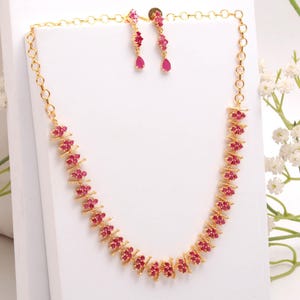 Pode incluir: Um conjunto de colar e brincos dourados. O colar e os brincos apresentam pequenas pedras preciosas vermelhas agrupadas. O colar tem um design de corrente delicado. Os brincos têm formato de gota.