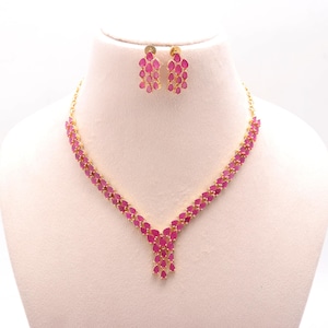 Könnte beinhalten: Ein goldfarbenes Halsketten- und Ohrring-Set. Die Halskette und die Ohrringe weisen ein Muster aus rosa tropfenförmigen Steinen auf. Die Halskette hat ein V-förmiges Design mit einem kaskadierenden Anhänger. Die Ohrringe sind eine Ansammlung derselben rosa Steine.
