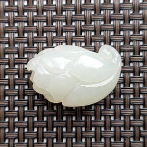 May include: A carved, off-white jade pendant in the shape of a flower bud. The pendant is smooth and polished, with detailed petal carvings. The background is a brown woven pattern.