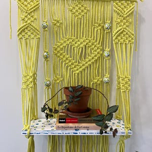 May include: A handmade macrame wall shelf in a bright yellow color. The shelf features a wooden dowel at the top, intricate knotting patterns, and a patterned shelf. Decorative ceramic beads and a small potted plant with books complete the design.