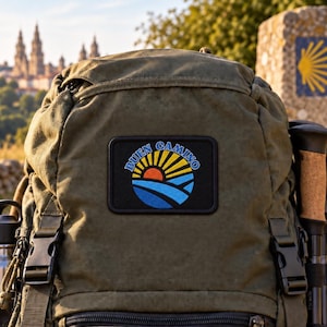 May include: An olive green backpack featuring a rectangular embroidered patch. The patch displays a stylised sun rising over blue hills with the text BUEN CAMINO arched above in blue letters. The backpack has black plastic buckles and straps.