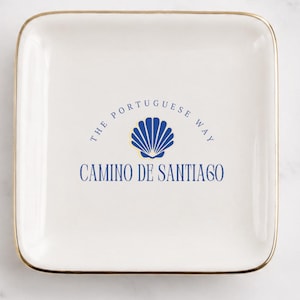 May include: A square white ceramic trinket dish with a gold-toned rim. The center features a blue scallop shell graphic with the text THE PORTUGUESE WAY arched above and CAMINO DE SANTIAGO printed in blue lettering below the shell icon.