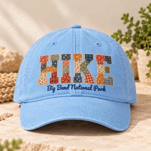 Op de afbeelding: Een lichtblauwe baseballpet met het woord HIKE in grote blokletters gevuld met een veelkleurig patchwork bloemen- en ruitpatroon. Onder het woord HIKE staat de tekst Big Bend National Park in donkerblauw cursief schrift.
