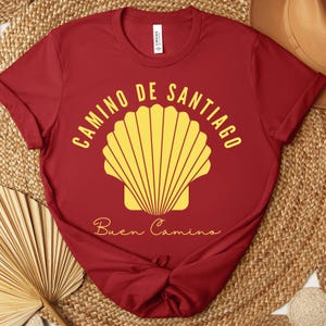 May include: A maroon t-shirt with the words "CAMINO DE SANTIAGO" in yellow, arched above a yellow scallop shell design. Below the shell, the words "Buen Camino" are written in cursive. The shirt is made of a soft material.